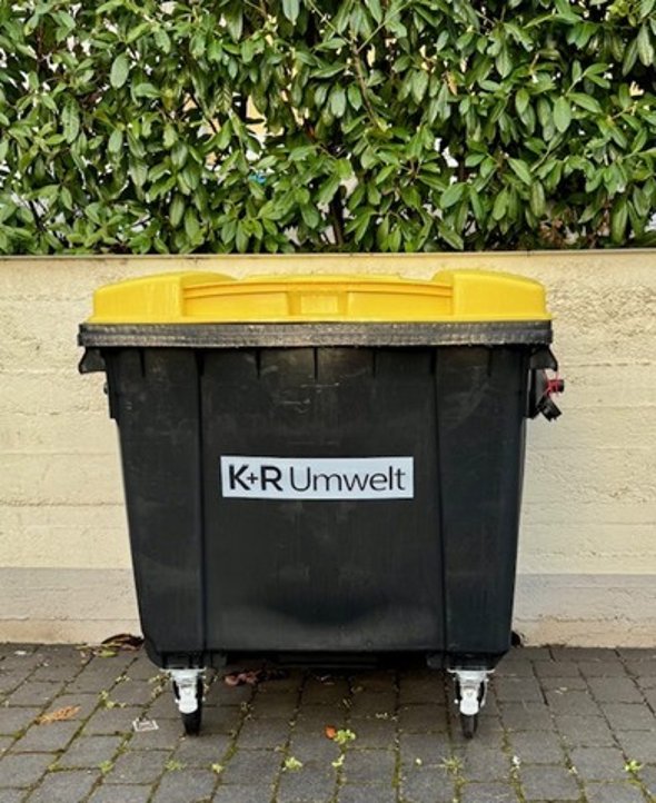 Vor einer Mauer, die unten einen grauen Streifen hat und darüber beige ist, stehet eine große Mülltonne. Sie ist schwarz und hat einen gelben rechteckigen Deckel. Auf der Tonne ist ein weißes Rechteck, in dem in schwarz "K+R Umwelt" steht. Über der Mauer ist eine Hecke mit grünen ovalen Blättern.