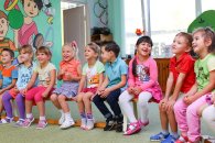 Auf eine Sportbank sitzen nebeneinander drei Jungen und sechs Mädchen im Kindergartenalter und lachen. Im Hintergrund ist eine Zimmertür, ein großes gekipptes Fenster und eine Wand, die mit einem Jungen und einem Mädchen mit Ball und Gymnastikband und Reifen im Sportdress bemalt ist.