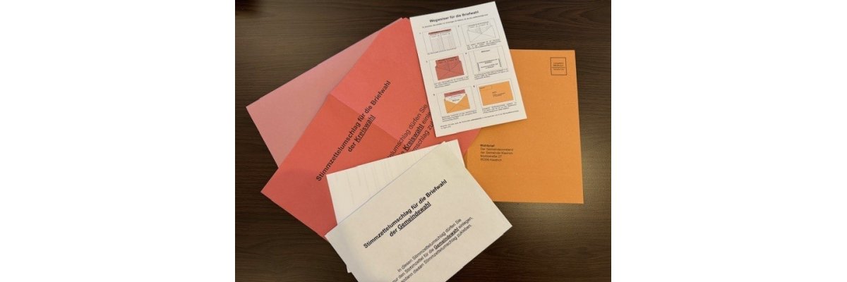 Auf einem schwarzen Tisch liegen mehrere Umschläge. Sie sind orange, pink und rosa. Darauf liegt ein weißer Umschlag mit schwarzer Schrift und ein weißer Zettel, auf dem erklärt wird, wie die Umschläge zu sortieren sind. 