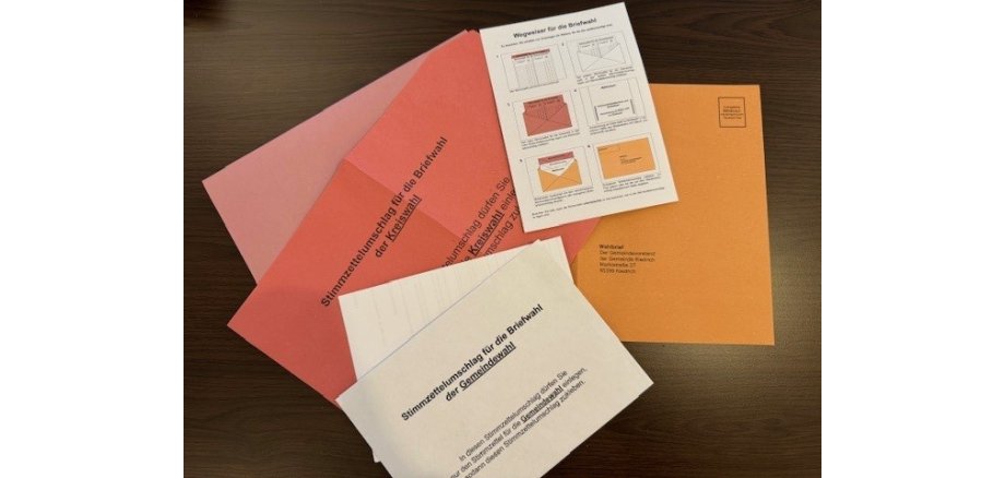 Auf einem schwarzen Tisch liegen mehrere Umschläge. Sie sind orange, pink und rosa. Darauf liegt ein weißer Umschlag mit schwarzer Schrift und ein weißer Zettel, auf dem erklärt wird, wie die Umschläge zu sortieren sind. 