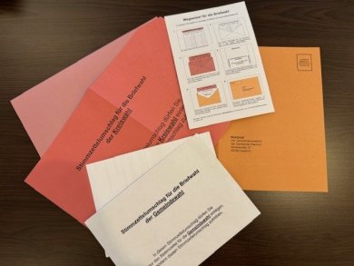 Auf einem schwarzen Tisch liegen mehrere Umschläge. Sie sind orange, pink und rosa. Darauf liegt ein weißer Umschlag mit schwarzer Schrift und ein weißer Zettel, auf dem erklärt wird, wie die Umschläge zu sortieren sind. 