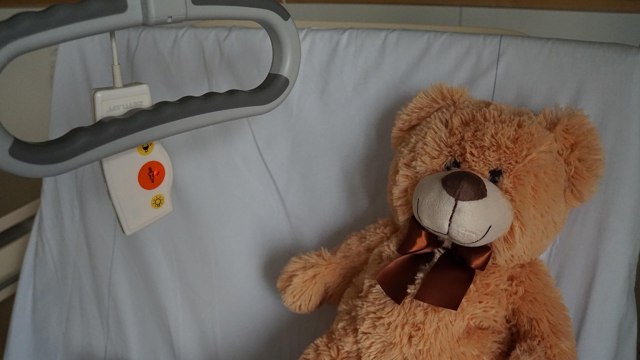 Man sieht einen Auschnitt eines Krankenbetts, mit einer aufgestellten Rückenlehne, in dem ein brauner Teddy mit einer braunen Halsbandschleife liegt. Neben ihm ist ein Notfallknopf und ein Haltegriff zum Hochziehen.