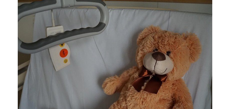 Man sieht einen Auschnitt eines Krankenbetts, mit einer aufgestellten Rückenlehne, in dem ein brauner Teddy mit einer braunen Halsbandschleife liegt. Neben ihm ist ein Notfallknopf und ein Haltegriff zum Hochziehen.