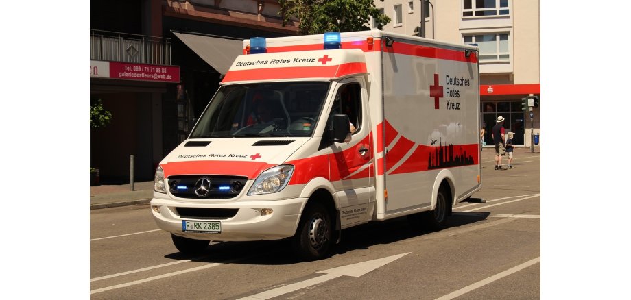 Auf einer Straße fährt ein Krankenwagen des Deutschen Roten Kreuzes mit Blaulicht. Im Hintergrund, Bürgersteig, ein Balkon, eine graue Markise, eine Sparkasse, eine Ampel und ein Mann und ein Junge in kurzer Hose und T-Shirt mit Sonnenhut und Sonnenmütze.