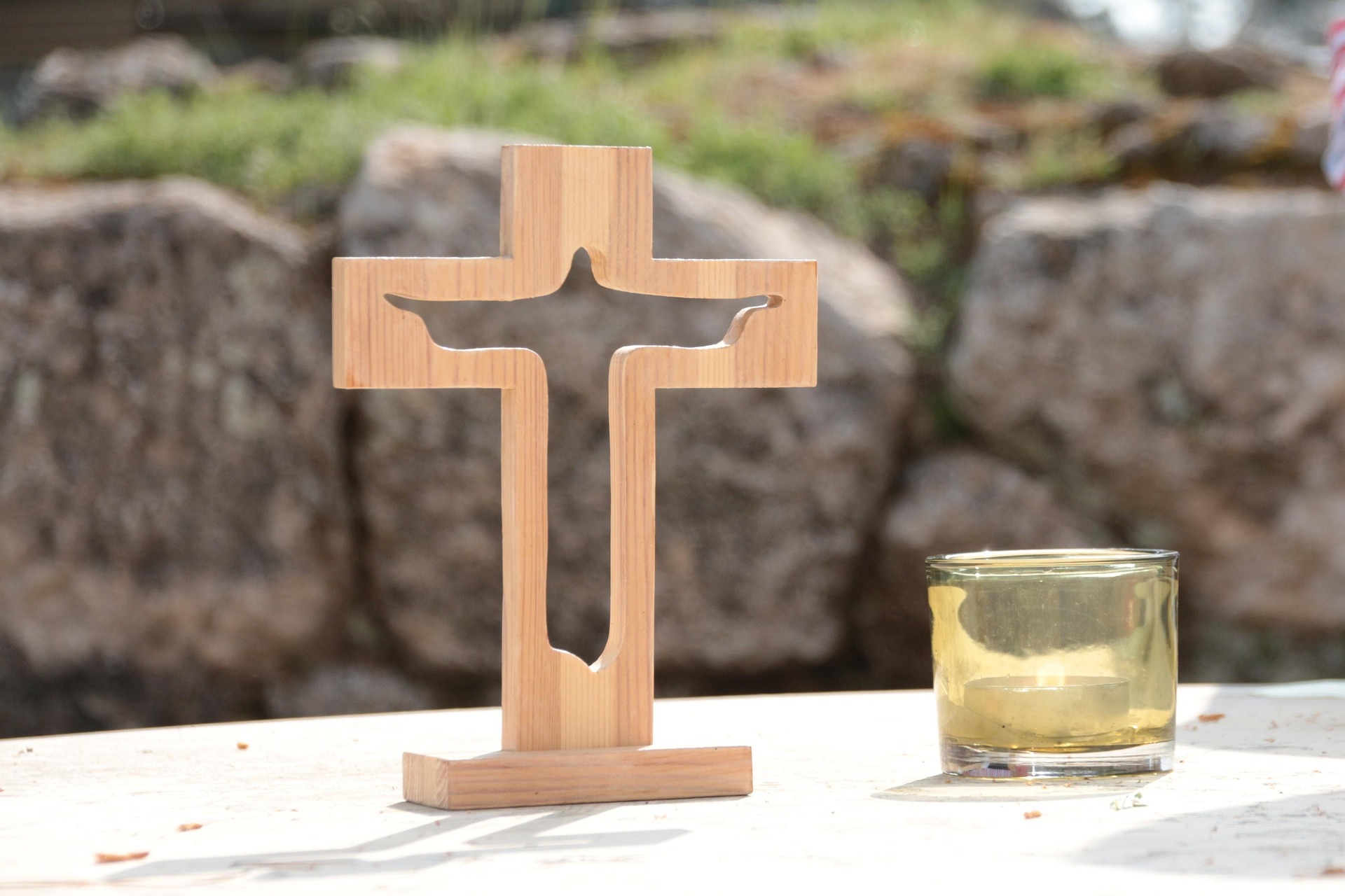 Auf einer weißen Tischdecke steht ein Kreuz aus Holz, in dem in der Mitte die Umrisse eines Engels sind. Daneben steht ein Teelicht mit einer Kerze. Im Hintergrund sind große aufeinander gestapelte Steine und eine Moosfläche.