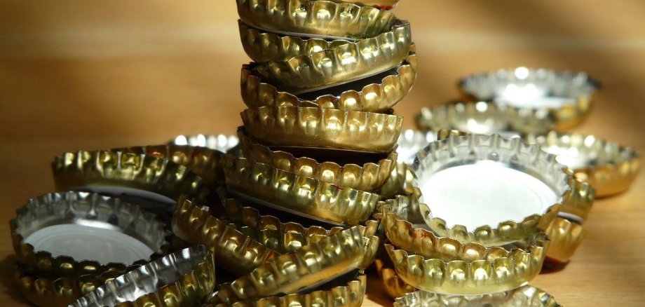 Auf einer braunen Holzfläche liegen goldene Kronkorken, quer übereinander und 14 Kronkorken wuren aufeinander gestapelt.