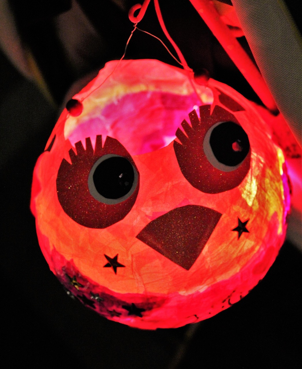 Vor einem dunklem Hintergrund ist eine runde rötliche Laterne an einem Stab, die leuchtet. Sie ist verziert mit zwei großen Augen mit Wimpern und einem Schnabel und daneben zwei Sternen.