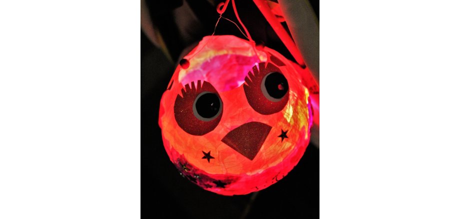 Vor einem dunklem Hintergrund ist eine runde rötliche Laterne an einem Stab, die leuchtet. Sie ist verziert mit zwei großen Augen mit Wimpern und einem Schnabel und daneben zwei Sternen.