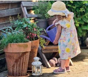 Marleen Ein kleines Mädchen im Blumenkleid und beigem Sonnenhut gießt mit einer blauen Gießkann zwei terrakottafarbene große Blumentöpfe, die mit Blumen bepflanzt sind, Davor steht eine weiße Laterne und im Hintergrund ein Holzstuhl, auf dem Blumentöpfe stehen und im Hintergrund eine belaubte Hecke.