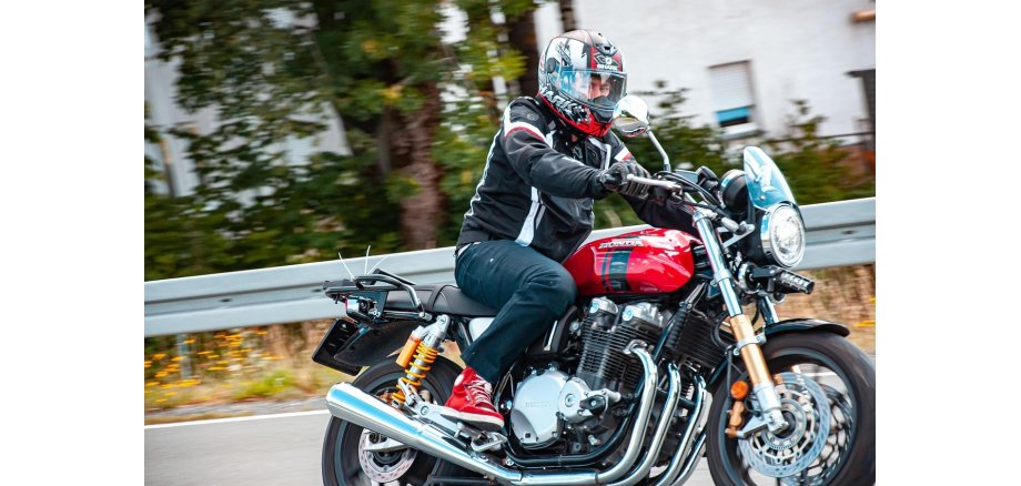 Auf einer Straße mit Leitplanke, hinter der verschwommen Bäume und Häuser sind, fährt ein Mann mit schwarzer Motorradjacke, die weiße und rote Streifen hat auf einer roten Honda. Er trägt eine dunkle Jeans, schwarze Handschuhe, rote Schuhe und einen Motorradhelm, der schwarz-rot-grau ist.