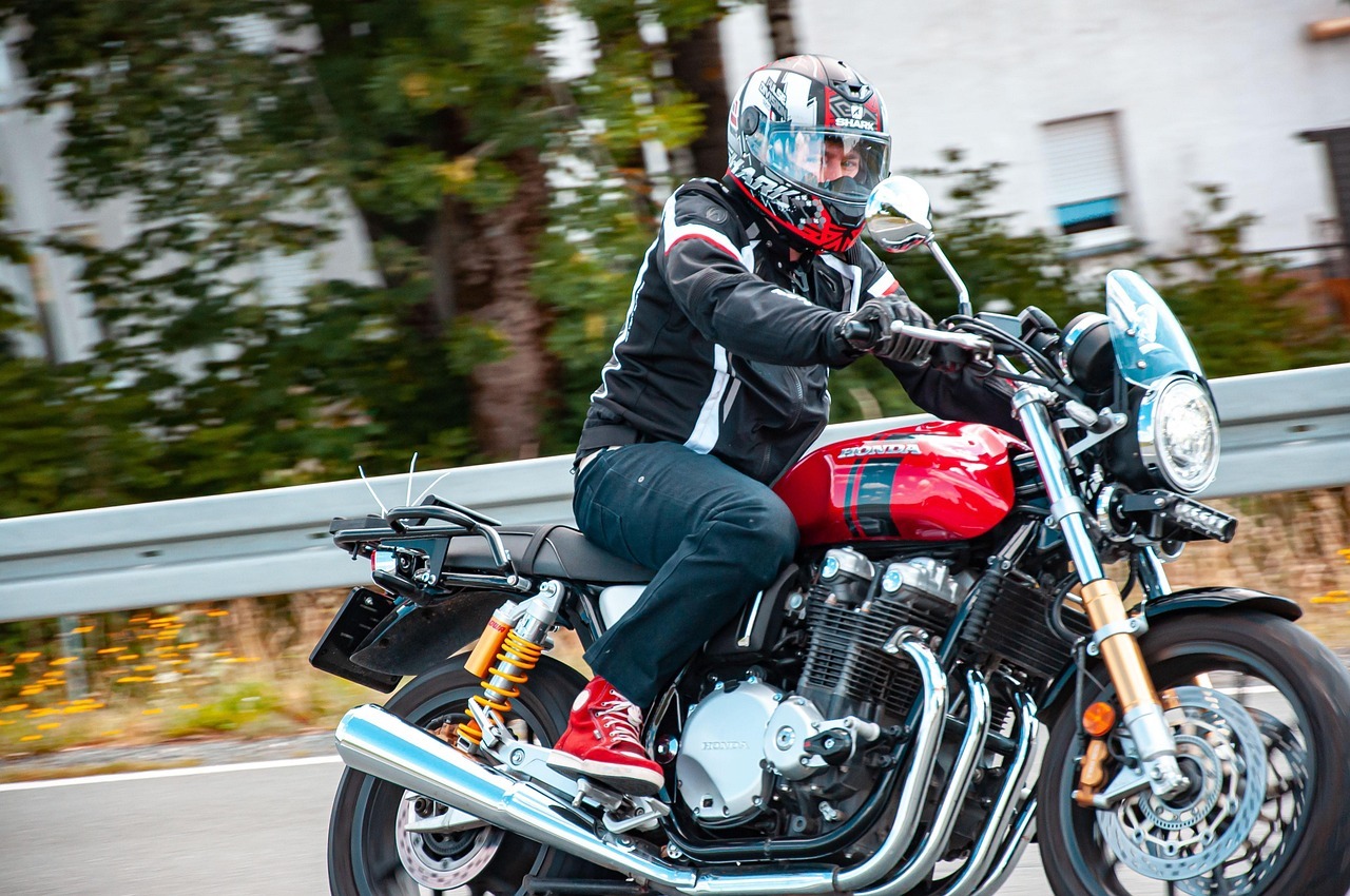 Auf einer Straße mit Leitplanke, hinter der verschwommen Bäume und Häuser sind, fährt ein Mann mit schwarzer Motorradjacke, die weiße und rote Streifen hat auf einer roten Honda. Er trägt eine dunkle Jeans, schwarze Handschuhe, rote Schuhe und einen Motorradhelm, der schwarz-rot-grau ist.