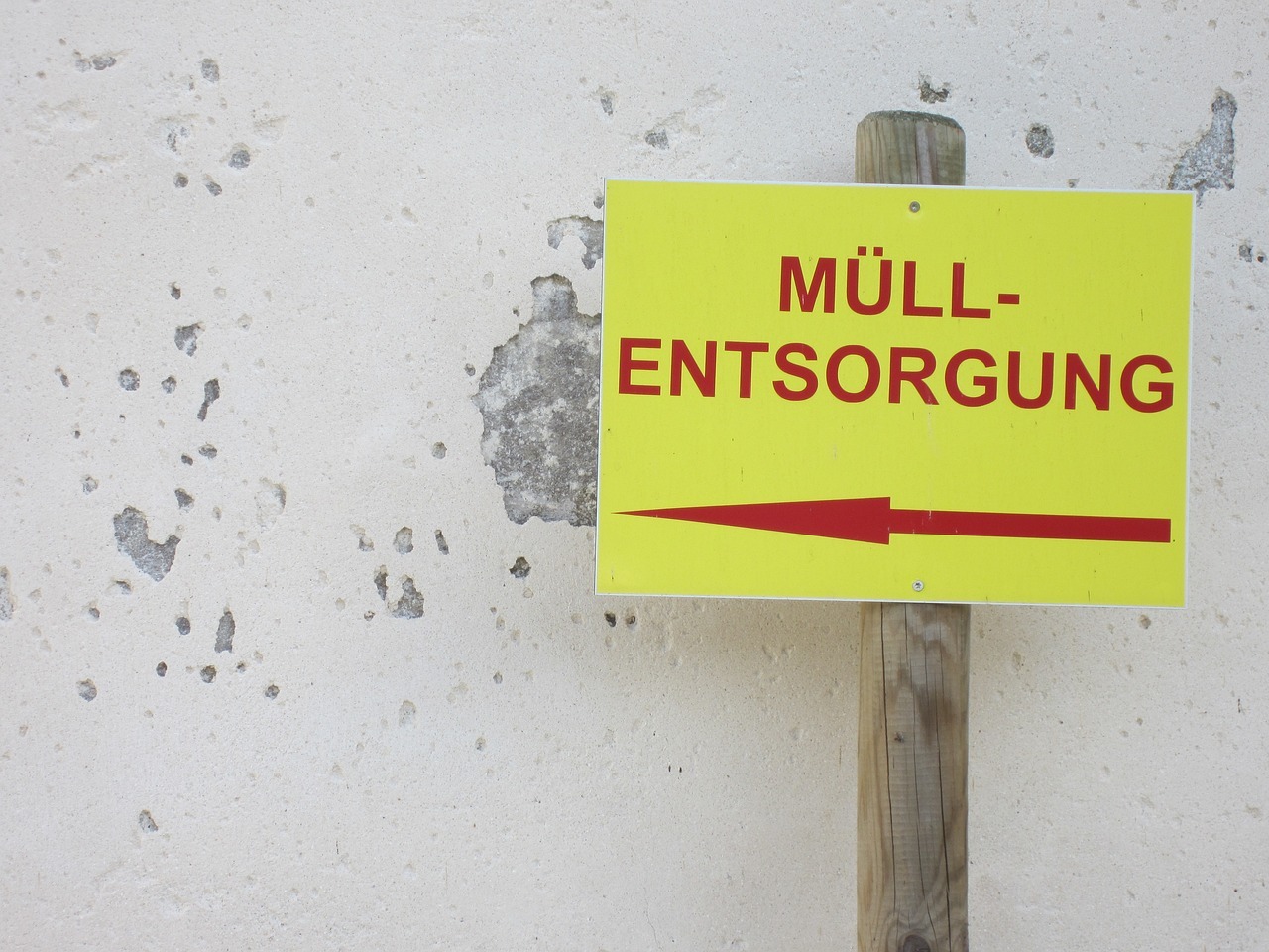 Vor einer weißen Wand, an der bereits der Putz abgeblättet ist und der Graue Beton durchschimmert, steht ein Holzpfahl, an dem ein Schild in gelber Neonfarbe angebracht ist. Auf dem Schild steht in roten Buchstaben "Müllentsorgung"  und ein Pfeil nach links