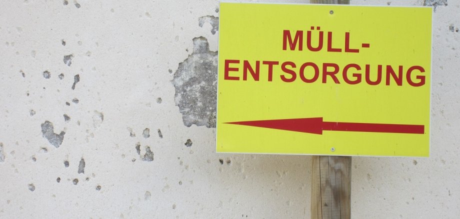 Vor einer weißen Wand, an der bereits der Putz abgeblättet ist und der Graue Beton durchschimmert, steht ein Holzpfahl, an dem ein Schild in gelber Neonfarbe angebracht ist. Auf dem Schild steht in roten Buchstaben "Müllentsorgung"  und ein Pfeil nach links