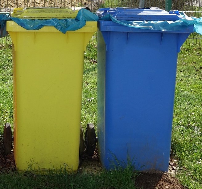 Auf einer Wiese steht eine gelbe Tonne und rechts davon eine blaue Tonne. Beide Tonnen haben blaue Müllsäcke in sich.