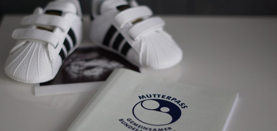 Auf einer weißen oberfläche liegt ein weisser Mutterpass mit dunkler Schrift, darüber ein Ultraschallbild und schwarz-weiße Baby-Sport-Sneaker.
