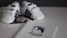 Auf einer weißen oberfläche liegt ein weisser Mutterpass mit dunkler Schrift, darüber ein Ultraschallbild und schwarz-weiße Baby-Sport-Sneaker.