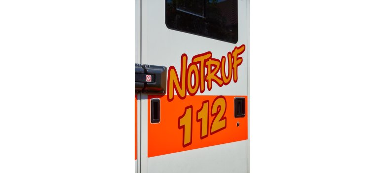 Auf der Tür eines Krankenwagens steht in moderner Schrift in orange, mit roter Umrandung, "Notruf" und auf einem nenorangenem Streifen, die Zahlen "112".