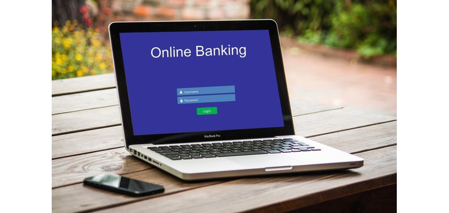 Auf einem Holztisch, der draußen steht, liegt ein schwarzes Handy und daneben steht ein aufgeklappter, silberner Laptop. Auf dem blauem Bildschirm steht "Online Banking" und die Login-Felder. Im Hintergrund sind verschwommen Blumen und ein Weg zu erkennen.
