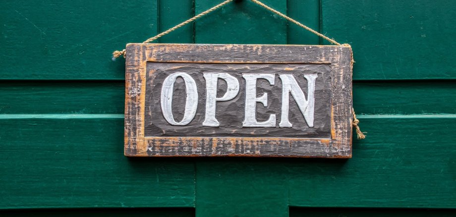 Schild geöffnet Auf dem Ausschnitt einer grünen Holztür mit Muster, hängt ein schraffiertes rechteckiges Holzschild an einer Kordel, die an einem runden silbernen Haken hängt. Auf dem Schild steht in weißen Buchstaben "OPEN".