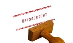 auf einem weißen Hintergrund liegt ein umgekippter rechteckiger Holzstempel.  Oberhalb ist in roten Buchstaben das Wort "Ortsgericht" gestempelt und ober- und unterhalb befinden sich jeweils ein dicker und ein dünner Strich in rot- und weiß. 