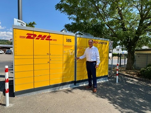 Ein Mann in dunkler Hose und weißem Hemd und einer Brille, steht vor einer gelben DHL-Packstation. Links und rechts stehen rot-weiße Pfosten und rechts ist ein großer, grünbelaubter Baum.