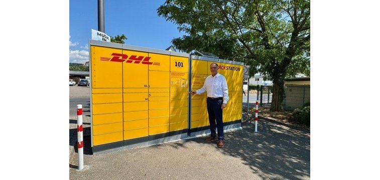 Ein Mann in dunkler Hose und weißem Hemd und einer Brille, steht vor einer gelben DHL-Packstation. Links und rechts stehen rot-weiße Pfosten und rechts ist ein großer, grünbelaubter Baum.