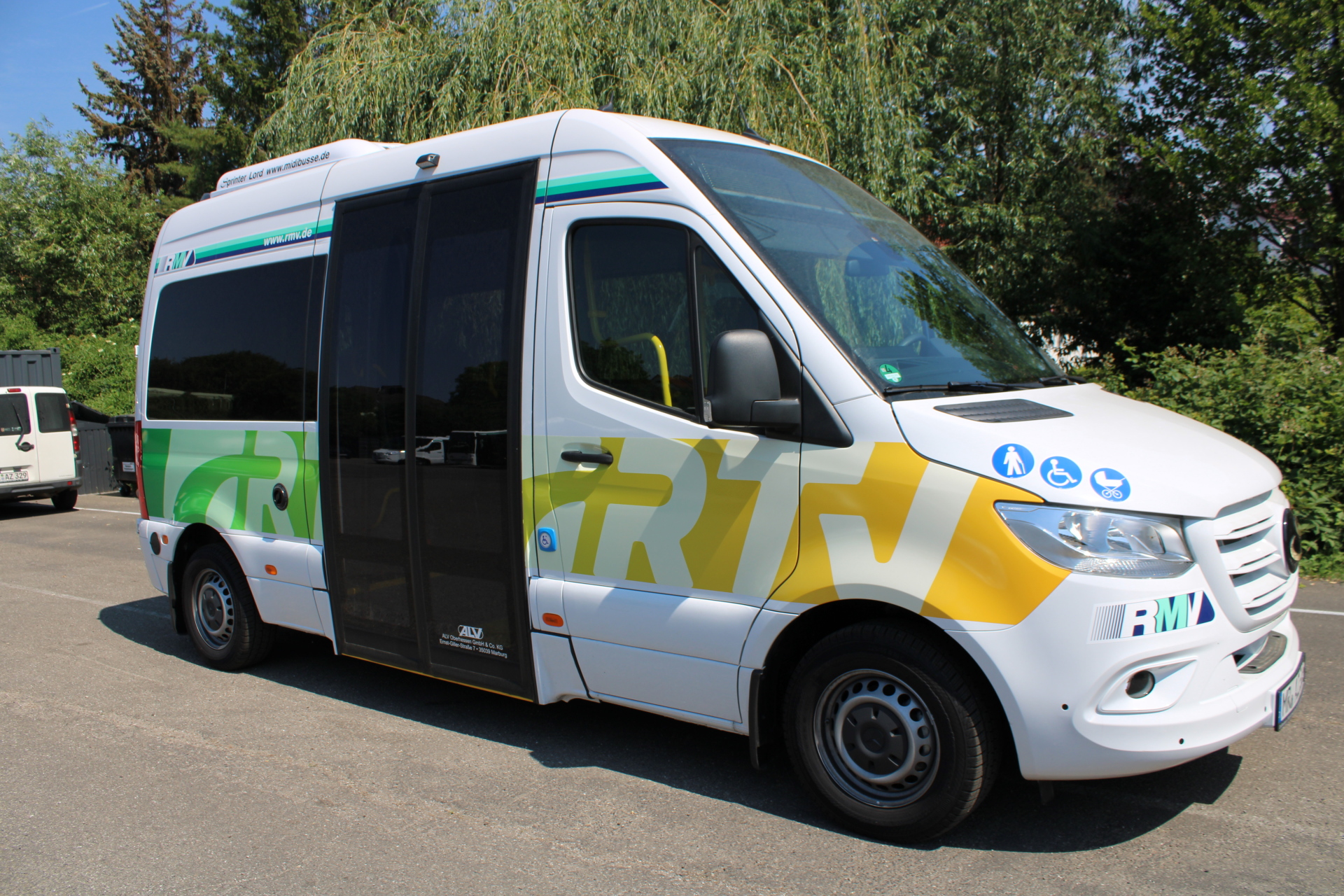 Auf einer Straße steht ein Rufbus vor mehreren Laubbäumen. Der Bus ist weiß und hat das Logo RTV in grün und gelb auf der Seite. Hinter der BEifahrertür ist eine breite Schiebetür mit schwarzen Scheiben bis auf den Boden. Auf der Motorhaube sind drei blaue Kreise mit weißen Symbolen- ein Mensch mit Stock, ein Mensch im Rollstuhl und ein Kinderwagen abgebildet. 