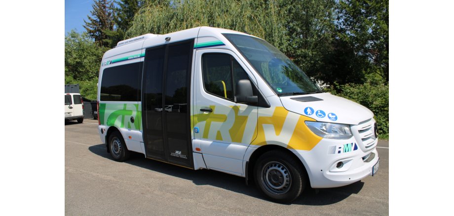 Auf einer Straße steht ein Rufbus vor mehreren Laubbäumen. Der Bus ist weiß und hat das Logo RTV in grün und gelb auf der Seite. Hinter der BEifahrertür ist eine breite Schiebetür mit schwarzen Scheiben bis auf den Boden. Auf der Motorhaube sind drei blaue Kreise mit weißen Symbolen- ein Mensch mit Stock, ein Mensch im Rollstuhl und ein Kinderwagen abgebildet. 