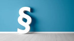 Satzung- Paragraphenzeichen Vor einer blauen wand, mit weißer Fußbodenleiste und grauem Fußboden mit Streifen, steht ein weißes Paragraphenzeichen an die Wand gelehnt.