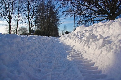 Ein Weg ist schneebedeckt und links und rechts ist Schnee aufgehäuft. Im Hintergrund sind schwarze Bäume zu erkennen. Der Himmel ist blau mit weißen Wolken.
