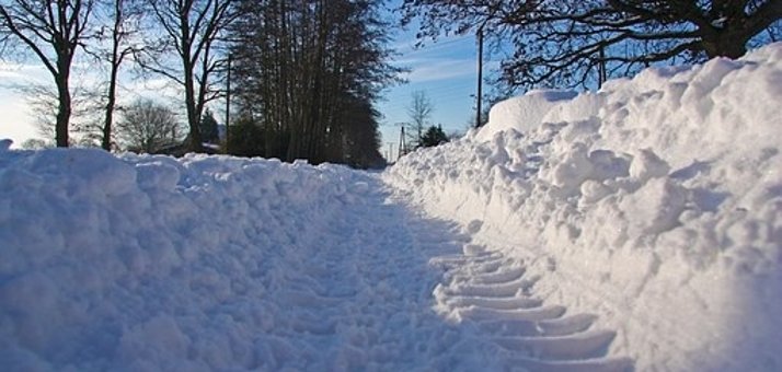 Ein Weg ist schneebedeckt und links und rechts ist Schnee aufgehäuft. Im Hintergrund sind schwarze Bäume zu erkennen. Der Himmel ist blau mit weißen Wolken.
