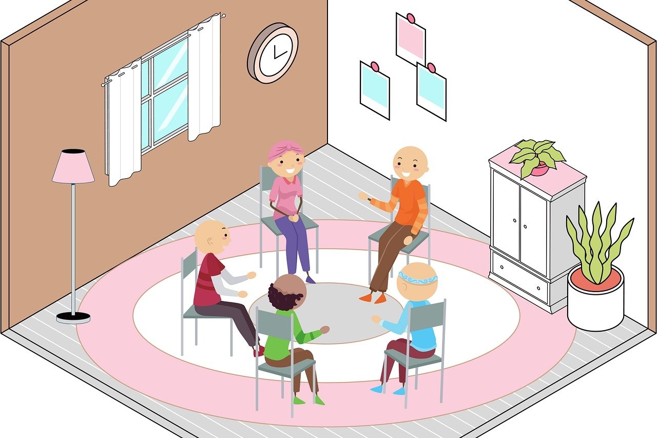 Das Bild ist gezeichnet. In einem Raum, sitzen 5 Personen auf Stühlen, die auf einem runden, weiß, rosa, beigen Teppich stehen. An der einen Wand hängen drei Bilder und davor steht eine Kommode mit einer Grünpflanze, neben der eine weitere Grünpflanze steht. An der anderen Wand hängt eine Uhr und es befindet sich ein Fenster mit Vorhängen und davor steht eine Stehlampe mit rosanem Lampenschirm.