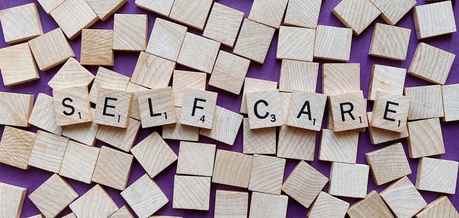 Auf einem lila Untergrund liegen mehrere Vierecke aus Holz durcheinanderverteilt. Auf diesen Quadraten liegen weitere acht Holzquadrate in der gleichen Größe, auf denen jeweils in schwarz ein Großbuchstabe und recht unten je eine kleine Zahl von 1 bis 4 steht. Die Buchstaben ergeben das Wort "SELF CARE".