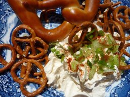 Auf einem weiß-blau-mit Blumen gemustertem Teller liegen ein Laugenbrezel und mehrere kleine Brezelchen sowie ein Klecks Spundekäse, der mit Frühlingszwiebelchen dekoriert ist.