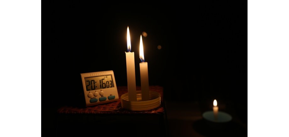 Auf einem geflochtenem Tisch steht ein digitaler weißer, rechteckiger Wecker und zeigt 20:16 03. Daneben stehen auf einem runden weißen Deckel zwei weiße brennende Kerzen und rechts daneben eine kleine weiße, brennende Kerze, die auf einem türkisfarbenem, runden Untersetzer steht.