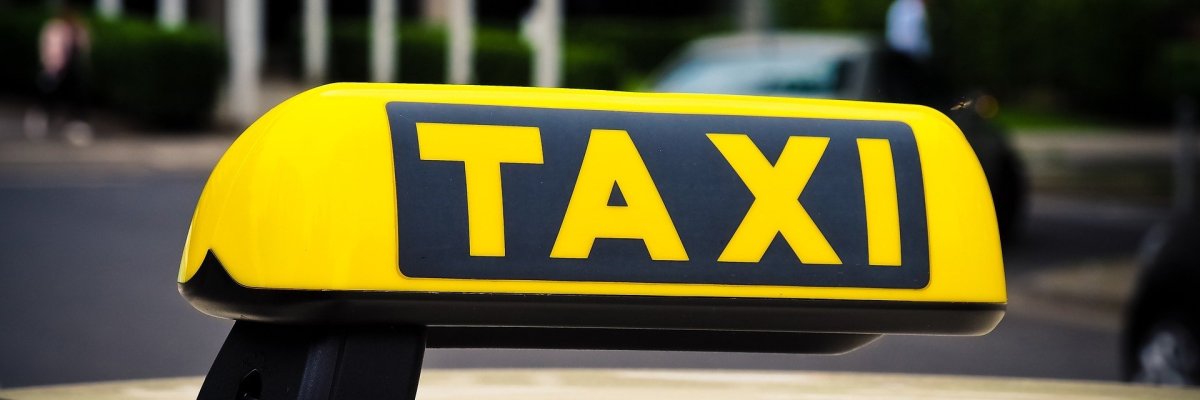 Das schwarz-gelbe Taxirechteck in dreiecksform auf dem Dach des beigen Taxis. Im Hintergrund sieht man verschwommen ein weißes Vordach, Hecken und eine Straße.