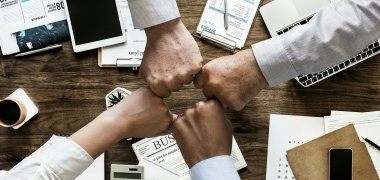 Teamwork Vier Fäuschte, die aus jeder Himmelsrichtung kommen und ein Viereck bilden. An den Armen ist eine langärmlige weiße Blusen und drei Hemdsärmel. Unten drunter sieht man einen Holztisch, auf dem ein Taschenrechner, ein Handy, ein Tablet, ein aufgeklappter Laptop, Zeitungen, Blöcke, Kugelschreibe, eine gefüllte Kaffeetasse stehen.
