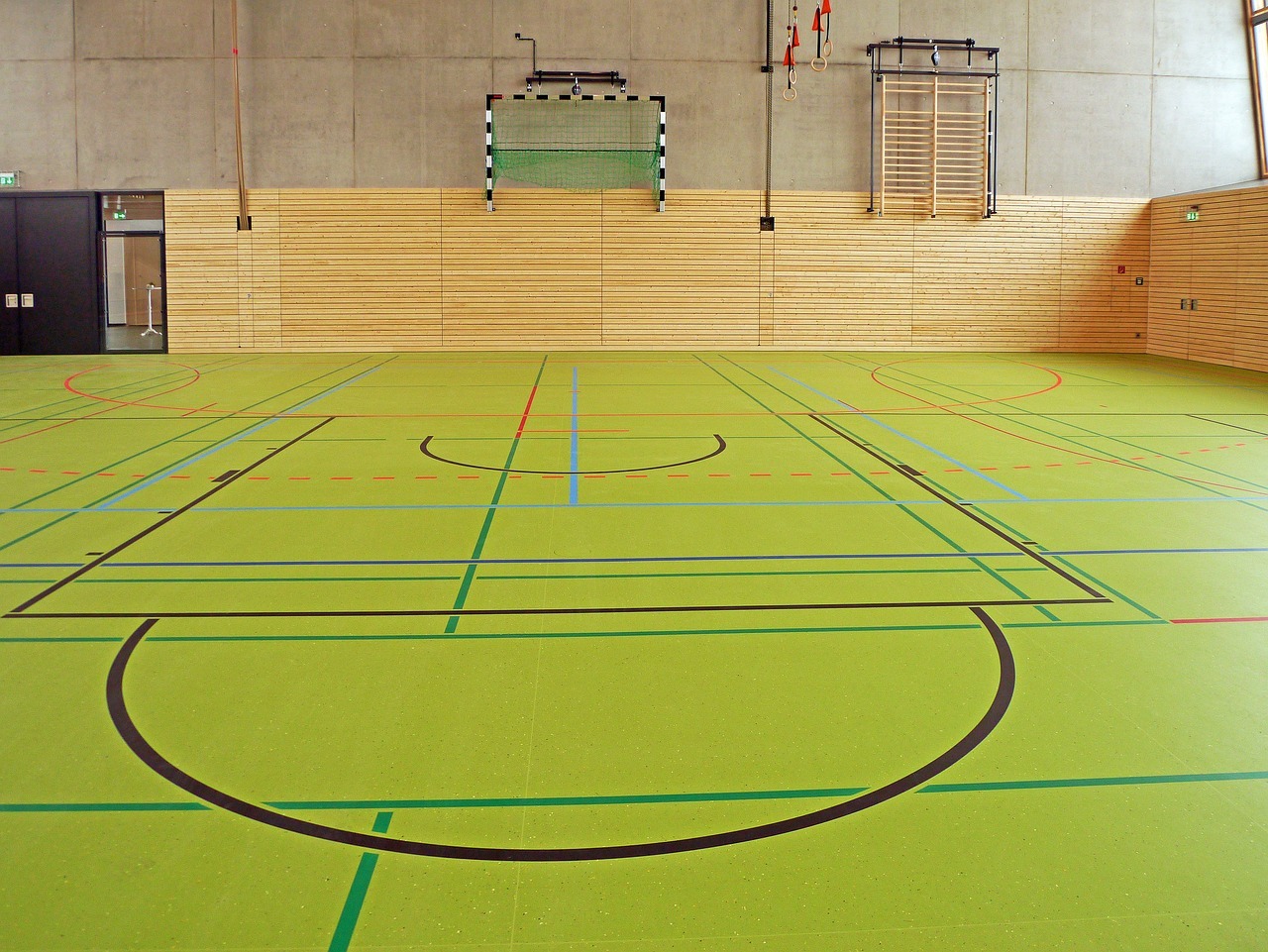 Eine Sporthalle mit einem grünen Hallenboden, auf den in verschiedenen Farben Felder eingezeichnet sind. An der Wand ist eine Holztäfelung und oben drüber Beton. An der Betonwand hängt ein grünes Fussballtor und eine Sprossenwand aus Holz.
