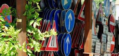 Unter einer Pergola, stehen verschiedenste Verkehrsschilder aufgereiht. Vorn ist um einen Pfosten eine Rankpflanze mit grünen Blättern.