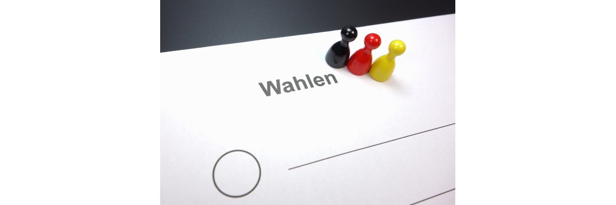 Ein weißer Zettel liegt auf einer schwarzen Fläche. Auf dem Zettel ist in schwarzen Buchstaben das Wort "Wahlen". Daneben steht ein schwarzes, ein rotes und ein gelbes Holzfigurenmännchen nebeneinander. Darunter sind zwei schwarze Striche und ein Kreis mit schwarz gemalt.
