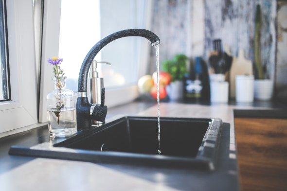 Ein rechteckiges Spülbecken in schwarz mit weißen Sprenkeln mit einem laufendem Wasserhahn in den gleichen Farben. Dahinter steht rechts vom Wasserhahn ein silbernes Gefäß zum Drücken mit Flüssigseife und links vom Wasserhahn einer durchsichtigen Vase mit einer lila Blüte. Dahinter befindet sich ein gekipptes Fenster und im L-Bereich der Arbeitsplatte ist verschwommen ein langer Kaktus, ein Schneidebrett, weiße Gefäße, rote Äpfel in einer Schale und grünes Basilikum in einem weißen Blumentopf zu sehen.