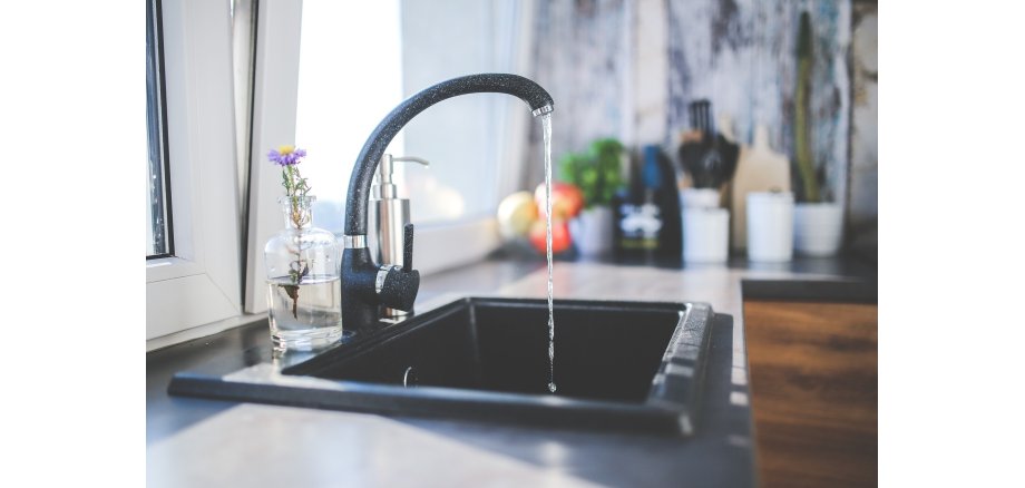 Ein rechteckiges Spülbecken in schwarz mit weißen Sprenkeln mit einem laufendem Wasserhahn in den gleichen Farben. Dahinter steht rechts vom Wasserhahn ein silbernes Gefäß zum Drücken mit Flüssigseife und links vom Wasserhahn einer durchsichtigen Vase mit einer lila Blüte. Dahinter befindet sich ein gekipptes Fenster und im L-Bereich der Arbeitsplatte ist verschwommen ein langer Kaktus, ein Schneidebrett, weiße Gefäße, rote Äpfel in einer Schale und grünes Basilikum in einem weißen Blumentopf zu sehen.