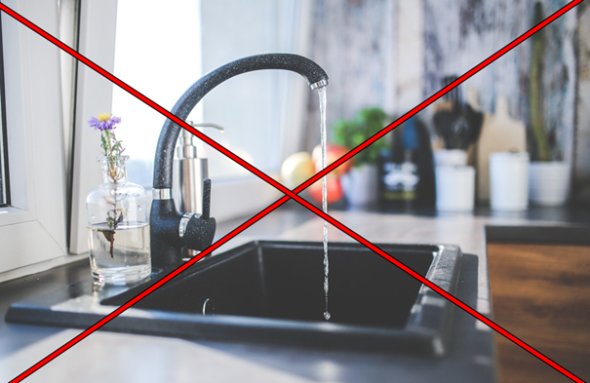 Ein rechteckiges Spülbecken in schwarz mit weißen Sprenkeln mit einem laufendem Wasserhahn in den gleichen Farben. Dahinter steht rechts vom Wasserhahn ein silbernes Gefäß zum Drücken mit Flüssigseife und links vom Wasserhahn einer durchsichtigen Vase mit einer lila Blüte. Dahinter befindet sich ein gekipptes Fenster und im L-Bereich der Arbeitsplatte ist verschwommen ein langer Kaktus, ein Schneidebrett, weiße Gefäße, rote Äpfel in einer Schale und grünes Basilikum in einem weißen Blumentopf zu sehen.