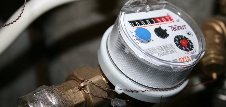 Metallleitung mit einer runden Wasseruhr aus weißem Kunststoff und durchsichtigem Deckel auf dem ein Strichcode, ein schwarzes Ziffernblatt mit den weißen Zahlen  von 0-9 und einem roten Zeiger und ein Rechteck mit fünf weiße Zahlenräder auf schwarzem HIntergrund und drei rote Zahlenräder auf rotem Hintergrund erkennbar ist. 