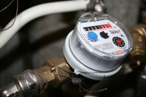Wasseruhr Metallleitung mit einer runden Wasseruhr aus weißem Kunststoff und durchsichtigem Deckel auf dem ein Strichcode, ein schwarzes Ziffernblatt mit den weißen Zahlen von 0-9 und einem roten Zeiger und ein Rechteck mit fünf weiße Zahlenräder auf schwarzem HIntergrund und drei rote Zahlenräder auf rotem Hintergrund erkennbar ist.