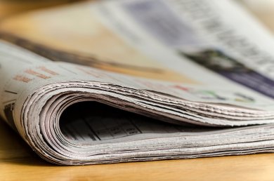 Zeitung Auf einem Holztisch liegt eine zusammengefaltete Zeitung. Die einzelnen Blätter im Vordergrund sind scharf, der Hintergrund ist verschwommen.