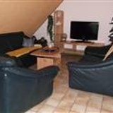 Wohnzimmer mit Dachschräge mit schwarzer Ledercouch mit drei Elementen, Holzcouchtisch, Zimmerpflanze und Fernseher auf einem braunen Fernsehtisch.