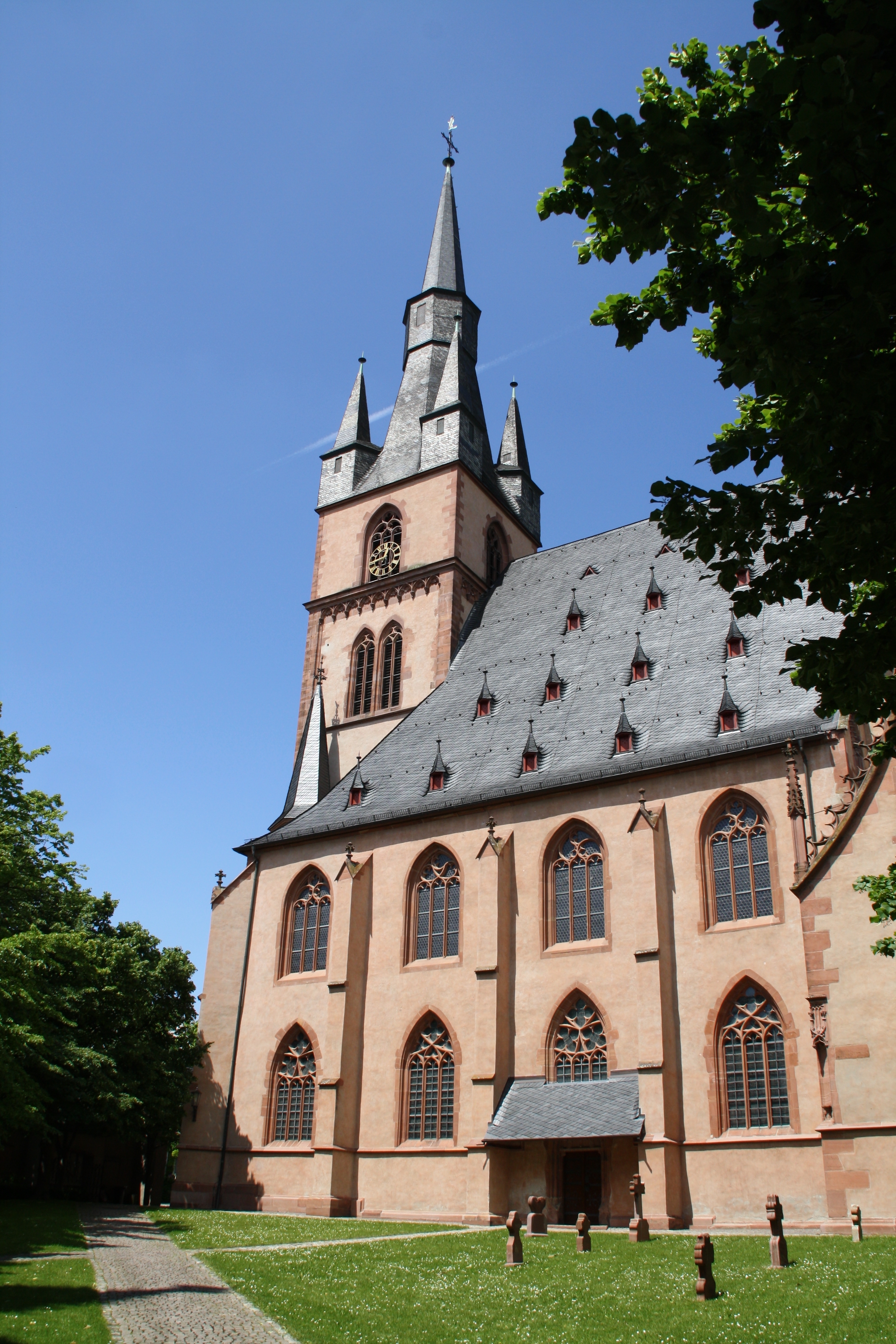 Blick auf die St. Valentinus und Dionysius Kiedrich mit dunklen Dachziegeln, rötlichen Steinen und dunkelroten Fensterumrandungen und Ziersteinen, dem Kirchturm mit einem großen und vier kleinen Türmen zwischen Bäumen und hinter einer Wiese mit einem Weg und sieben sandsteinfarbenen Grabsteinen vor einem blauen Himmel.