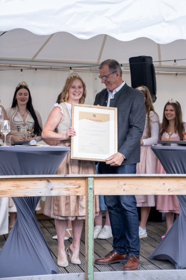 Die ehemalige Weinkönigin trägt ein weiß-beige-farbenes Dirndl und eine goldene Krone mit Gemeindewappen, Reben und Trauben auf dem Kopf. Rechts neben Ihr steht im grauem Trachten-Jacket, Bürgermeister Steinmacher. Beide halten einen großen Bilderrahmen mit goldenem Rahmen und einer Urkunde in der Hand. Im HIntergrund weitere Weinmajestäten.  Links und rechts davon stehen Bistrotische mit grauen Hussen.