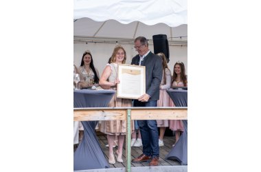 Die ehemalige Weinkönigin trägt ein weiß-beige-farbenes Dirndl und eine goldene Krone mit Gemeindewappen, Reben und Trauben auf dem Kopf. Rechts neben Ihr steht im grauem Trachten-Jacket, Bürgermeister Steinmacher. Beide halten einen großen Bilderrahmen mit goldenem Rahmen und einer Urkunde in der Hand. Im HIntergrund weitere Weinmajestäten. Links und rechts davon stehen Bistrotische mit grauen Hussen.